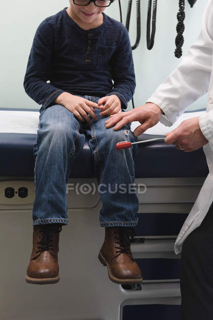 Vista frontal de un joven médico masculino asiático examinando a un niño caucásico paciente de rodilla en una clínica. Médico masculino usando martillo reflejo - foto de stock