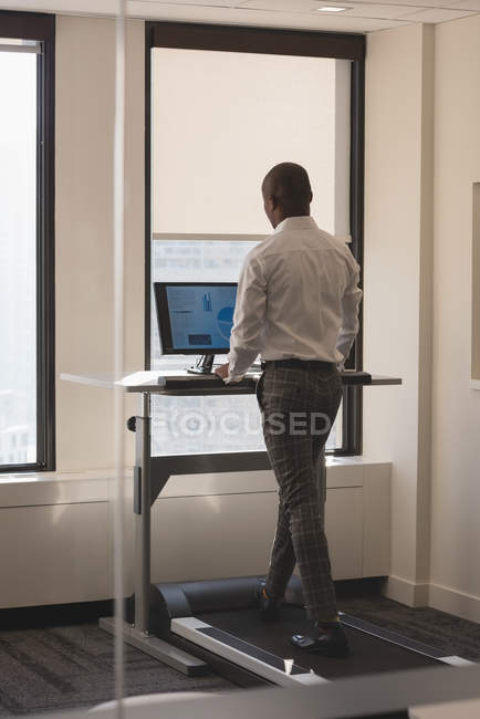 Vista lateral del hombre de negocios que trabaja mientras hace ejercicio en la cinta de correr en la oficina moderna - foto de stock
