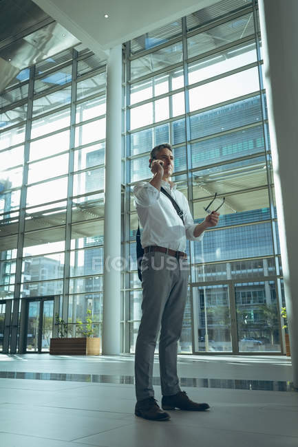 Vista lateral de un hombre de negocios confiado hablando en el teléfono móvil mientras sostiene sus gafas en una gran oficina vacía - foto de stock