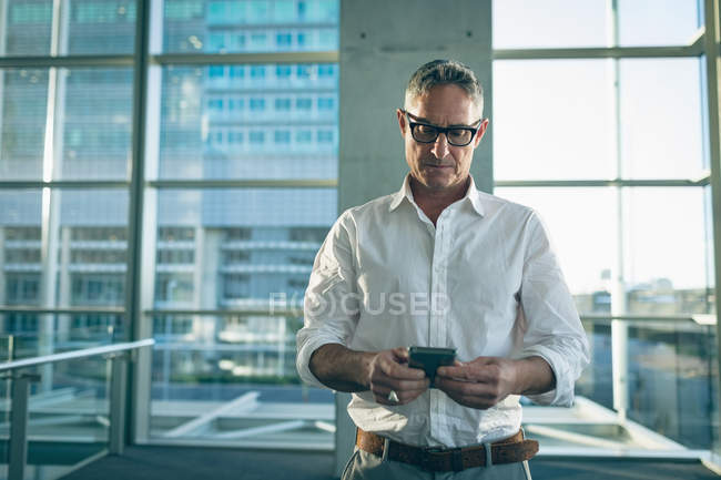 Вид спереди бизнесмена, смотрящего на свой мобильный телефон в офисе рядом с большими окнами, показывающими здание на заднем плане — стоковое фото