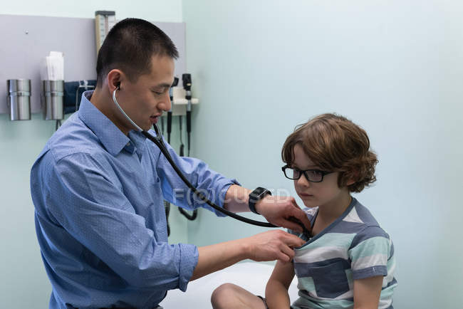 Vista laterale di giovane asiatico maschio medico esaminando caucasico ragazzo paziente con strumento in una clinica — Foto stock