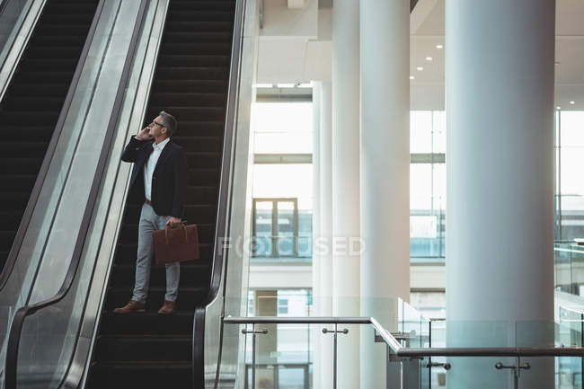 Дальний взгляд бизнесмена с портфелем разговаривающего по телефону на эскалаторе в офисе — стоковое фото