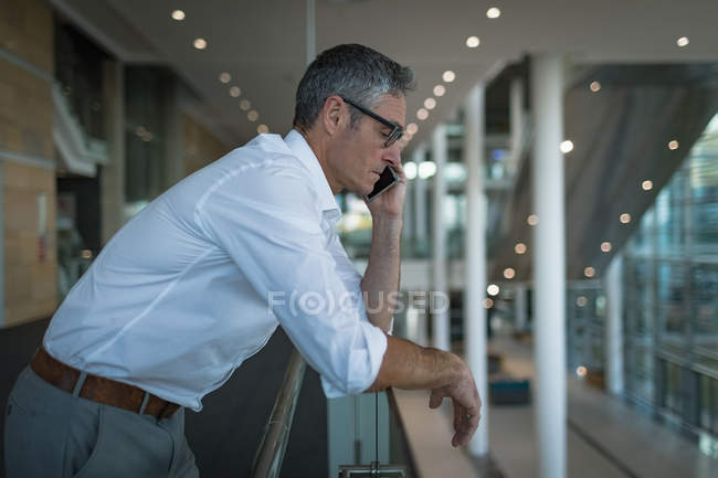 Vista lateral do empresário apoiando-se nos trilhos enquanto falava ao telefone no escritório — Fotografia de Stock