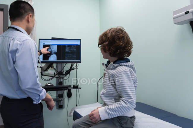 Vista trasera de joven asiático médico masculino mostrando a chico caucásico paciente de rayos X en la pantalla de la computadora - foto de stock
