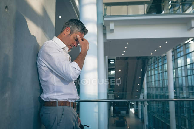 Side view of stressed businessman leaning against the wall while thinking and holding hie head in office — Stock Photo