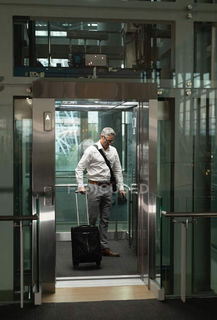 Вид спереди бизнесмена с дорожным чемоданом, смотрящего на команду в лифте в офисе — стоковое фото