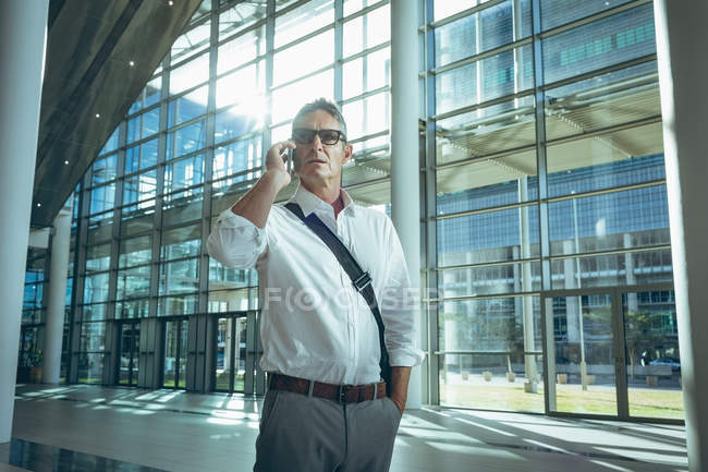 Front view of a tensed businessman talking on the mobile phone in a large empty office with buildings in the background — Stock Photo