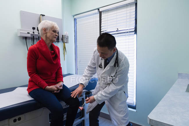 Молодой врач-азиат осматривает пожилого пациента рефлекторным молотком в клинике — стоковое фото