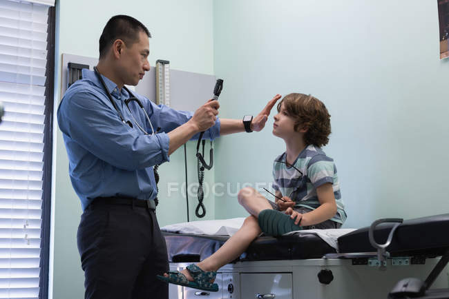 Vista laterale del giovane medico maschio asiatico che esamina un ragazzo caucasico paziente orecchio con strumento in una clinica — Foto stock