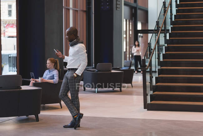 Vista lateral do empresário de pé usando telefone celular no lobby no escritório moderno — Fotografia de Stock