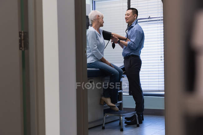 Vista lateral del médico masculino que comprueba la presión arterial del medidor de presión arterial del paciente mayor en la clínica - foto de stock