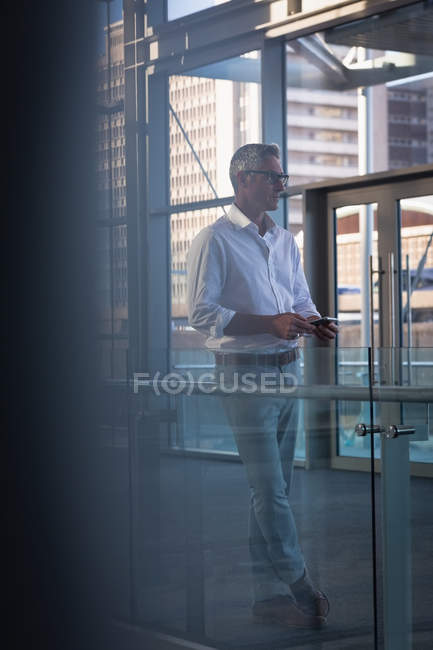 Vista lateral de um empresário atencioso com telefone celular olhando para longe ao lado de grandes janelas mostrando um prédio de escritórios em segundo plano — Fotografia de Stock