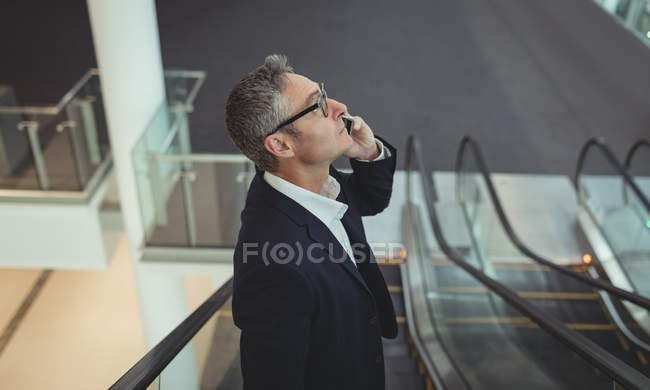 Vista lateral del hombre de negocios hablando en el teléfono móvil en la escalera mecánica en la oficina - foto de stock
