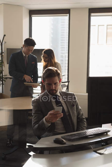 Vista lateral do empresário sentado usando telefone celular no escritório moderno com colegas de trabalho discutindo sobre tablet digital int he background — Fotografia de Stock