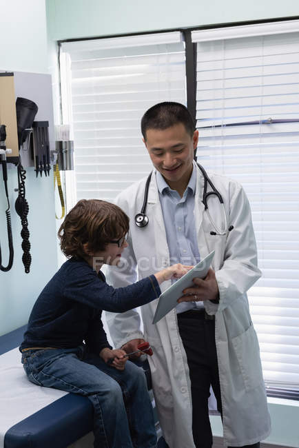 Vue de face du jeune médecin masculin asiatique et du patient garçon caucasien utilisant une tablette numérique à la clinique. Médecin masculin interagissant avec le patient — Photo de stock