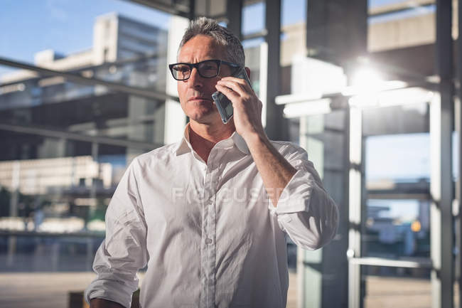 Close-up view of a businessman talking on the phone in the office against bright sunlight next to big windows showing the city in the background — Stock Photo