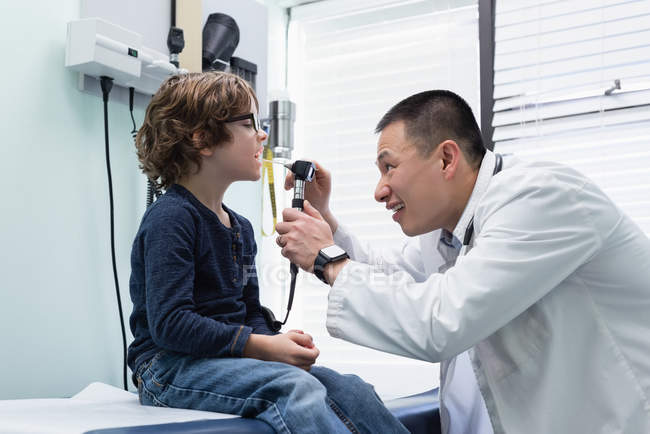 Vista laterale di giovane asiatico maschio medico esaminando caucasico ragazzo paziente con strumento in una clinica — Foto stock