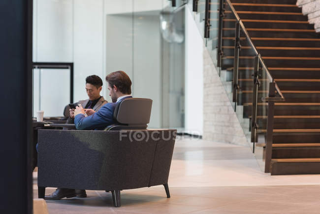 Vista lateral de empresários usando telefone celular sentado nas poltronas no lobby no escritório — Fotografia de Stock