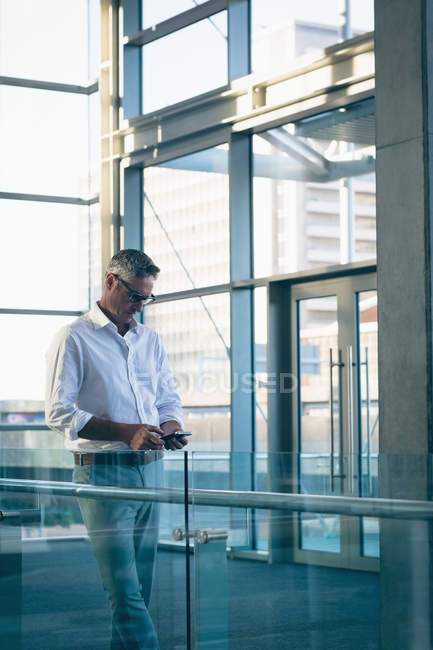 Vista laterale di un uomo d'affari concentrato che usa il suo cellulare nell'ufficio accanto a grandi finestre che mostrano un edificio per uffici sullo sfondo — Foto stock