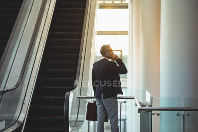 Rear view of businessman with briefcase talking on the phone near the escalator in office — Stock Photo