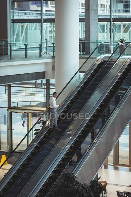 Vista elevada del hombre de negocios mirando su teléfono inteligente en la escalera mecánica en la oficina - foto de stock