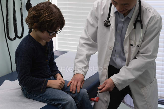 Visão frontal do jovem asiático médico masculino examinando caucasiano menino paciente joelho em uma clínica. Médico masculino usando o martelo reflexo — Fotografia de Stock