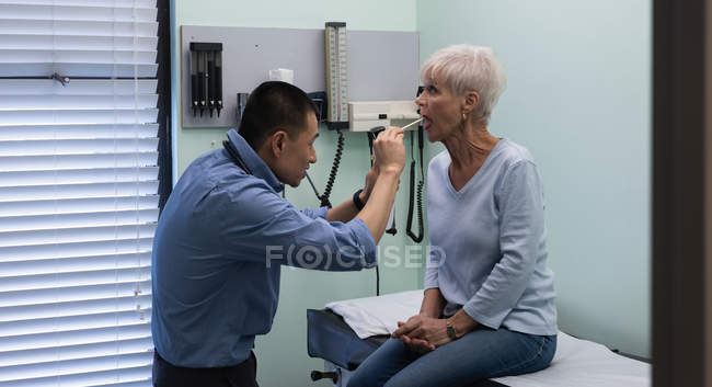 Vista laterale del giovane medico maschio asiatico che esamina un paziente anziano con torcia medica in clinica — Foto stock