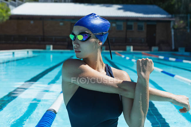 Vue de face d'une jeune nageuse avec des lunettes de bain qui s'étirent dans la piscine par une journée ensoleillée — Photo de stock