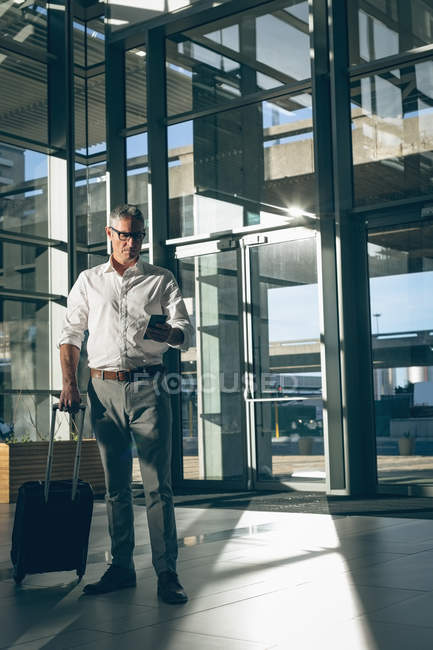 Vista frontal de un hombre de negocios sosteniendo una bolsa de viaje mientras revisa su teléfono móvil en la oficina - foto de stock