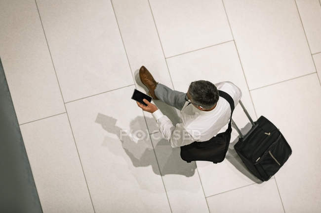 Elevated view of businessman with travel suitcase looking at his mobile phone while walking in the corridor — Stock Photo