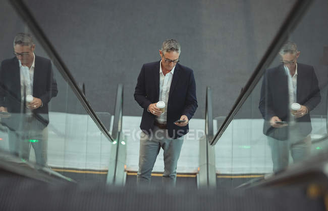 Blick auf Geschäftsmann mit Kaffeetasse, der sein Handy auf Rolltreppe im Büro benutzt — Stockfoto