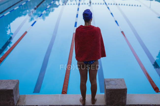 Vista posteriore del giovane nuotatore femminile avvolto in asciugamano in piedi vicino alla piscina — Foto stock