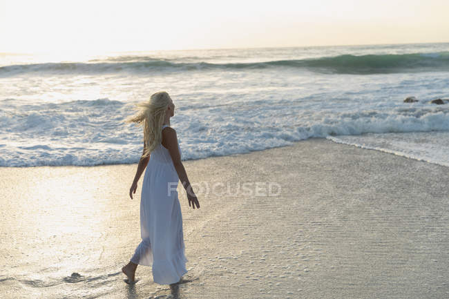 Вид сбоку на белокурую красивую женщину, идущую по пляжу в солнечный день. Она идет по морю — стоковое фото