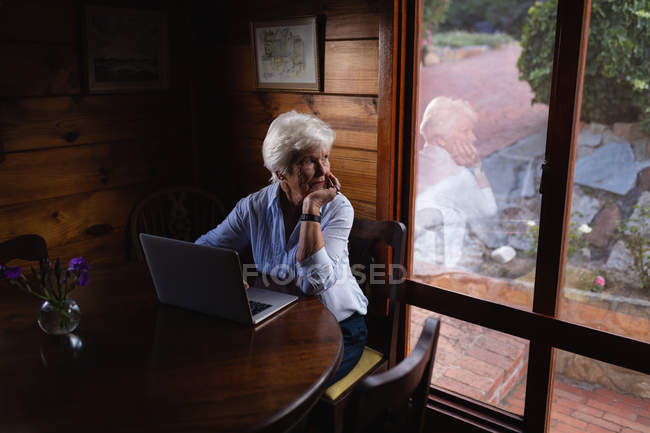 Вид спереди активной пожилой женщины, сидящей и работающей с ноутбуком, отводя взгляд от дома — стоковое фото