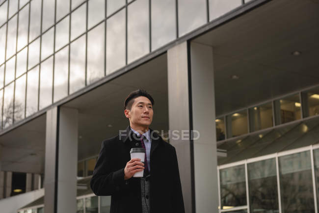 Front view of young Asian businessman with coffee cup walking on street in the city — Stock Photo