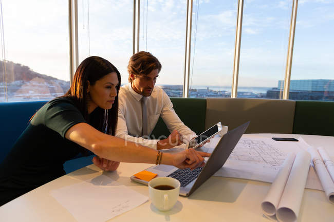 Vista lateral de jovens executivos sentados à mesa e trabalhando em laptop enquanto bebem uma xícara de café em um escritório moderno — Fotografia de Stock