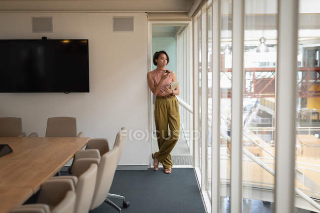 Vorderansicht einer asiatischen Geschäftsfrau, die mit ihrem Handy telefoniert und ein digitales Tablet in der Hand hält, während sie sich im Büro an ein Fenster lehnt — Stockfoto