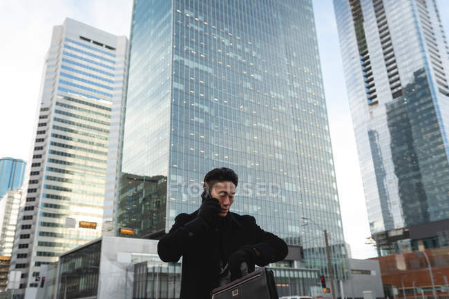 Visão de baixo ângulo do jovem empresário asiático olhando para smartwatch enquanto fala no telefone celular na rua na cidade — Fotografia de Stock