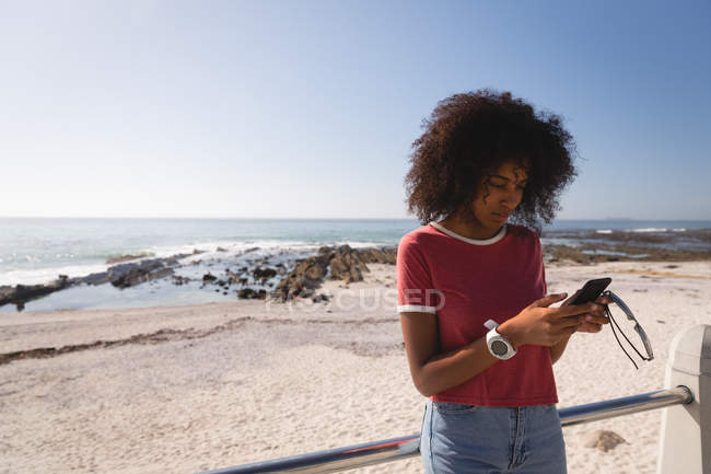 Vue de face de la belle femme afro-américaine utilisant un téléphone portable à la plage sur le soleil. Lunettes de maintien — Photo de stock