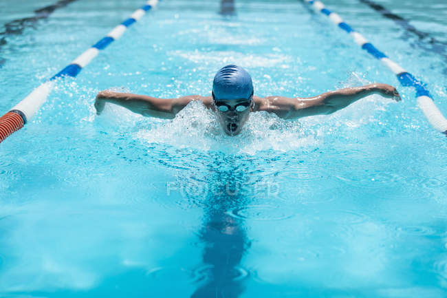 Front view of young Caucasian male swimmer in the middle of swimming butterfly stroke in outdoor swimming pool on sunny day — Stock Photo