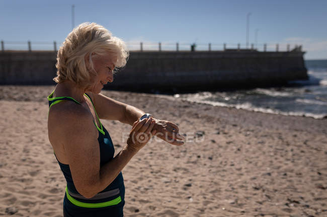 Vista lateral de uma mulher idosa ativa usando seu smartwatch na praia sob o sol — Fotografia de Stock