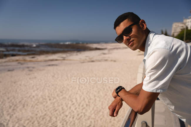 Вид сбоку на вдумчивого смешанного расового человека стоящего и смотрящего на камеру на пляже на солнце — стоковое фото