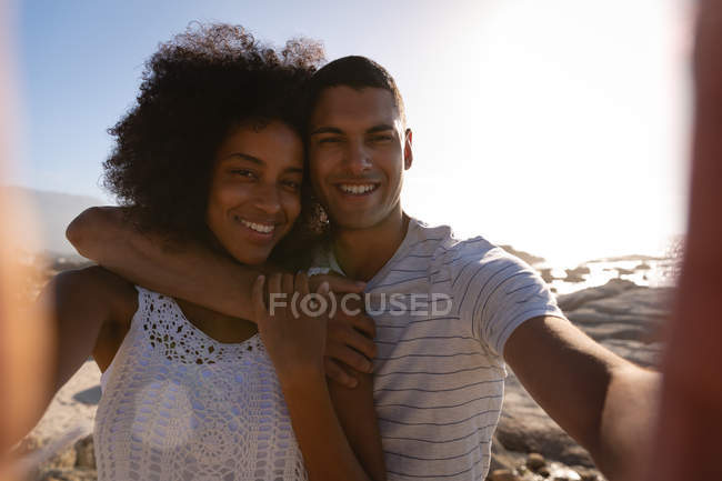 Vorderansicht eines afrikanisch-amerikanischen Paares, das lächelt und in die Kamera schaut, während es ein Selfie in der Nähe des Meeres macht — Stockfoto