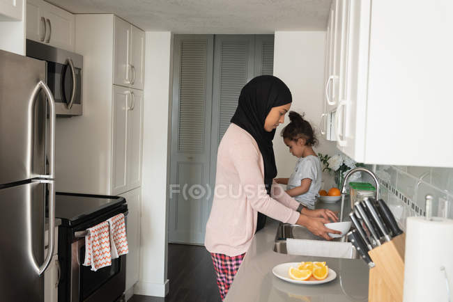 Vista lateral de la madre de raza mixta que usa hijab trabajando en la habitación de la cocina mientras su hija está sentada detrás de ella en casa - foto de stock
