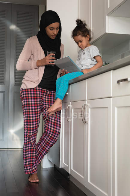 Vista de ángulo bajo de madre de raza mixta usando hijab e hija juntos usando tableta digital en la sala de cocina en casa - foto de stock