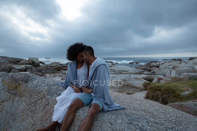 Vista frontal de la pareja afroamericana sentados y mirándose el uno al otro y relajándose cerca del lado del mar al atardecer - foto de stock