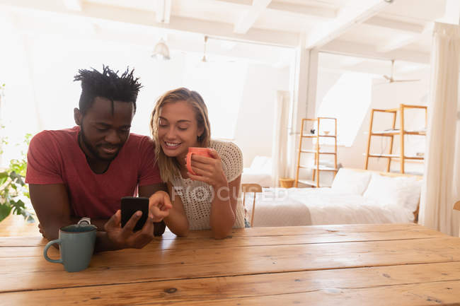 Vista frontal de sorrir e usar telefone celular em casa enquanto toma uma xícara de chá — Fotografia de Stock