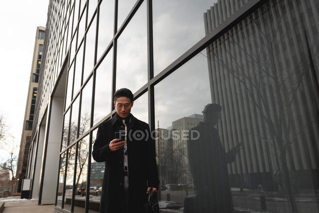Vista frontal de un joven empresario asiático bien vestido utilizando el teléfono móvil mientras pasea por un edificio moderno en la calle de la ciudad - foto de stock