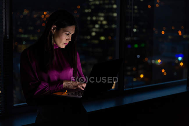 Вид спереди на молодую представительницу смешанной расы, работающую на ноутбуке возле окна в современном офисе — стоковое фото