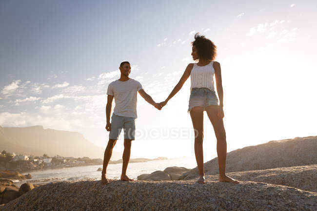 Low angle view of African-american couple holding hands and standing on rock near sea side on sunset — Stock Photo